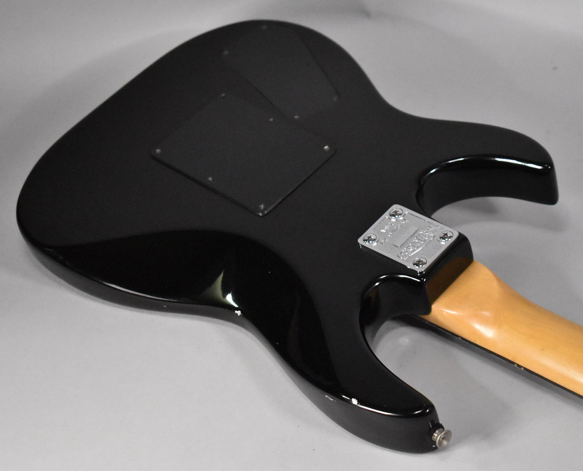 The back of a glossy black electric guitar, angled to show the neck joint, chrome neck plate, and rear tremolo cover.