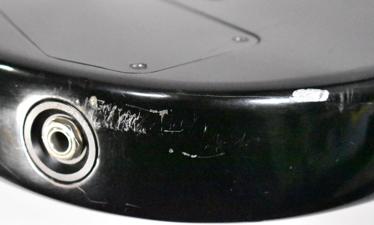 Close-up of the black guitar body's lower edge, showing the input jack, heavy scratching, and a large chip in the finish.