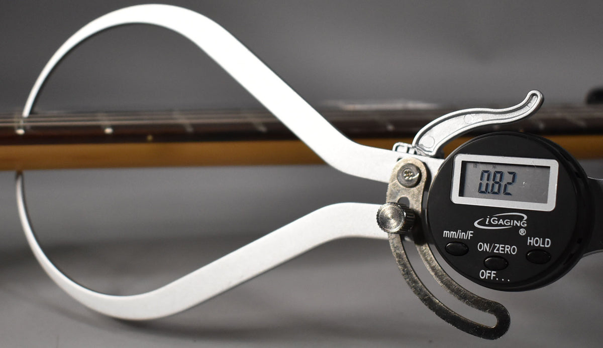 A digital caliper measures the string height above the dark fretboard of the guitar, displaying "0.82" on its screen.