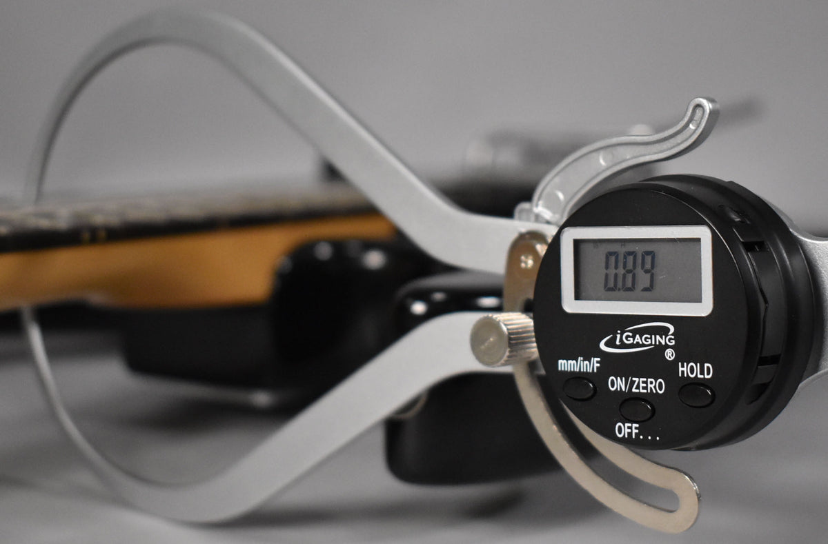 A digital caliper measures the black guitar's fret height at 0.89, showing the dark fretboard and light wood neck.