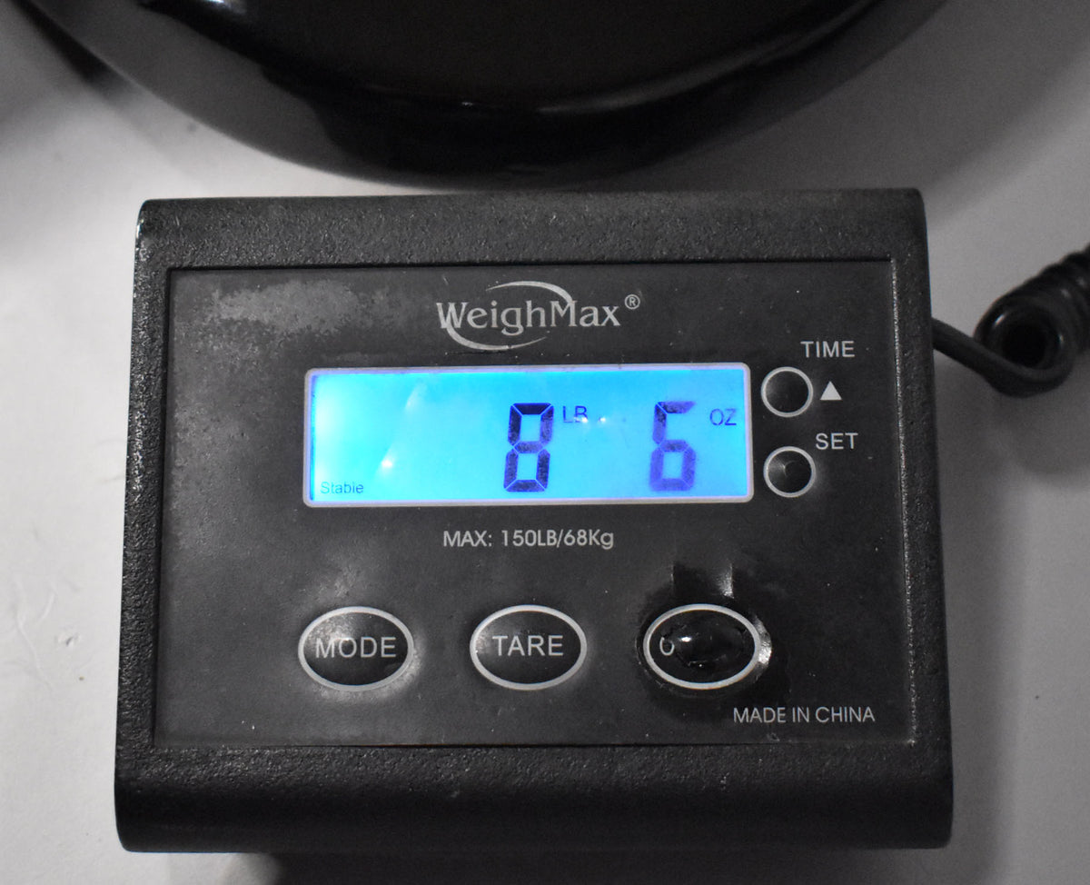 A close-up of a digital scale showing "8 LB 6 OZ" on its blue display, with the black guitar body partially visible above.