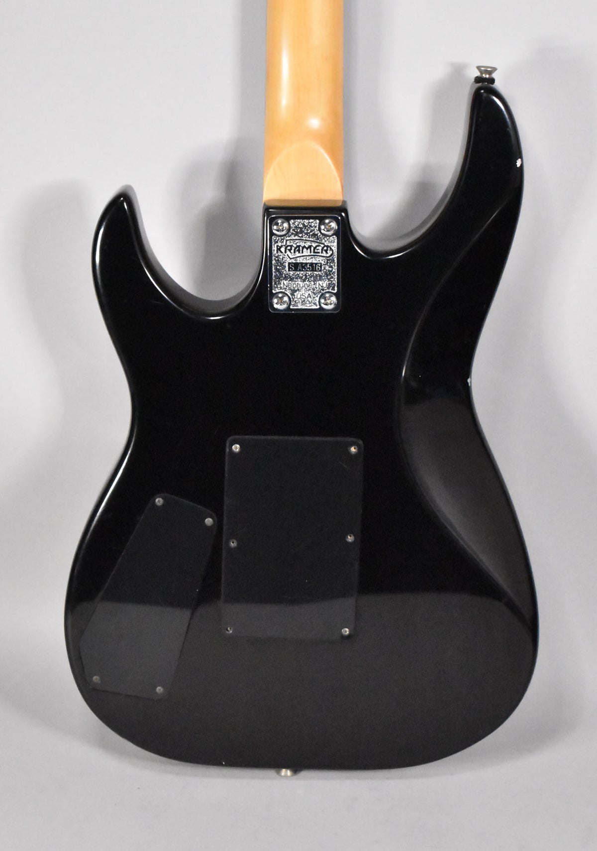The guitar's glossy black body is seen from the back, showing a natural finish neck, chrome neck plate, and two black control cavity covers.