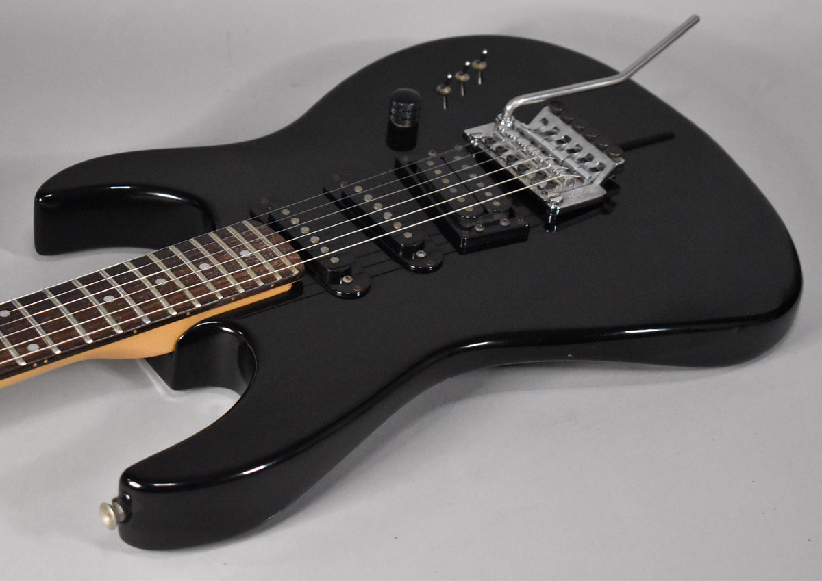 Gloss black electric guitar, angled from the lower bout, showing three humbucker pickups, a tremolo bridge, and three control knobs.