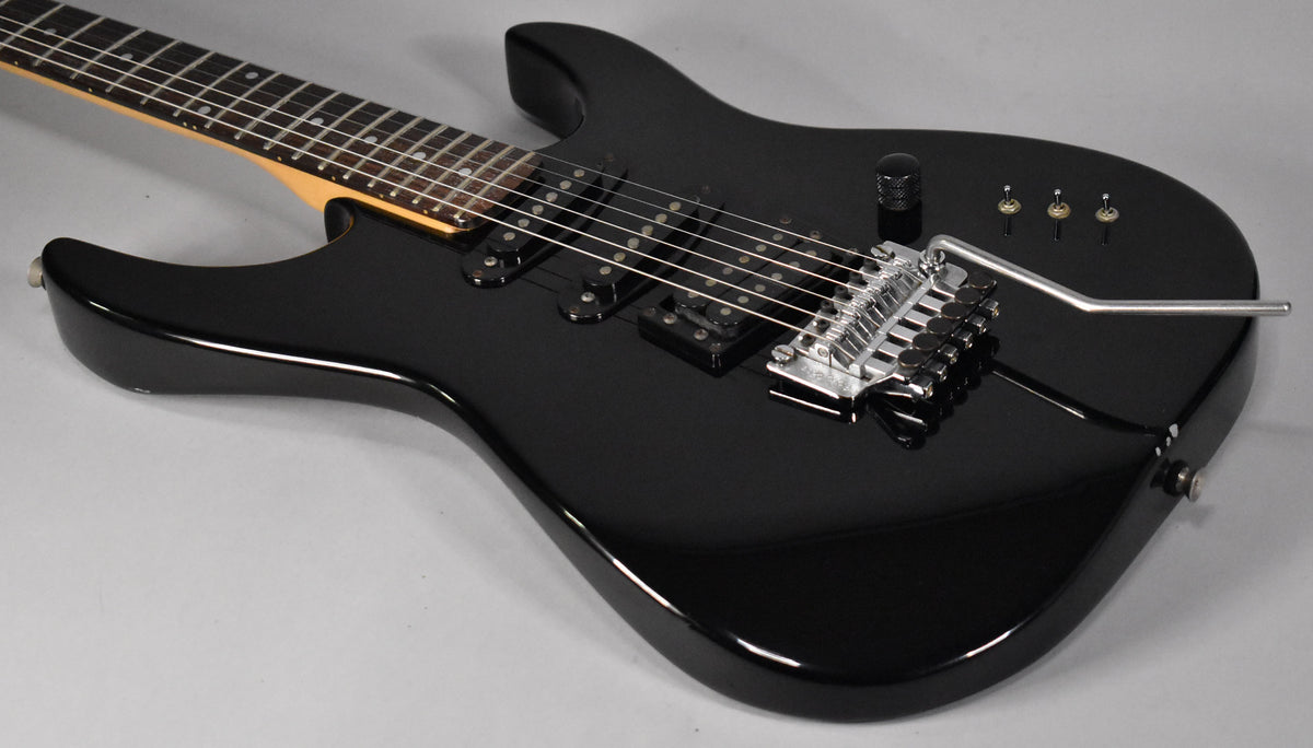 Black electric guitar, angled view of the body and lower neck, showing three humbucker pickups, a tremolo bridge, and three mini-toggle switches.