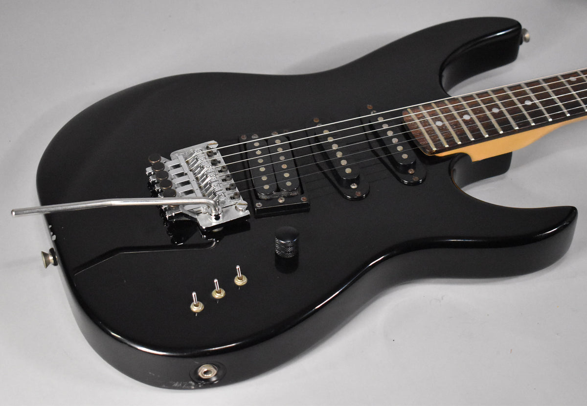 Black electric guitar at an angle, showing a Floyd Rose tremolo, a bridge humbucker, two single-coil pickups and three mini-toggle switches.