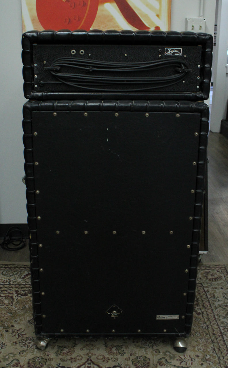 Rear view of the black tuck-and-roll amp head and cab, showing the head's two inputs and coiled cables, and the cab's casters.