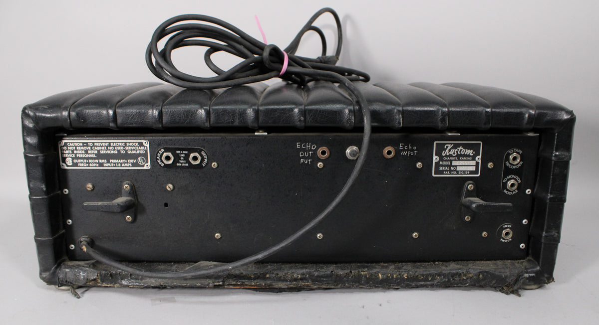 Rear view of the black tuck-and-roll amp head, showing various inputs, outputs, and a power cord coiled on top, with visible wear along the bottom edge.