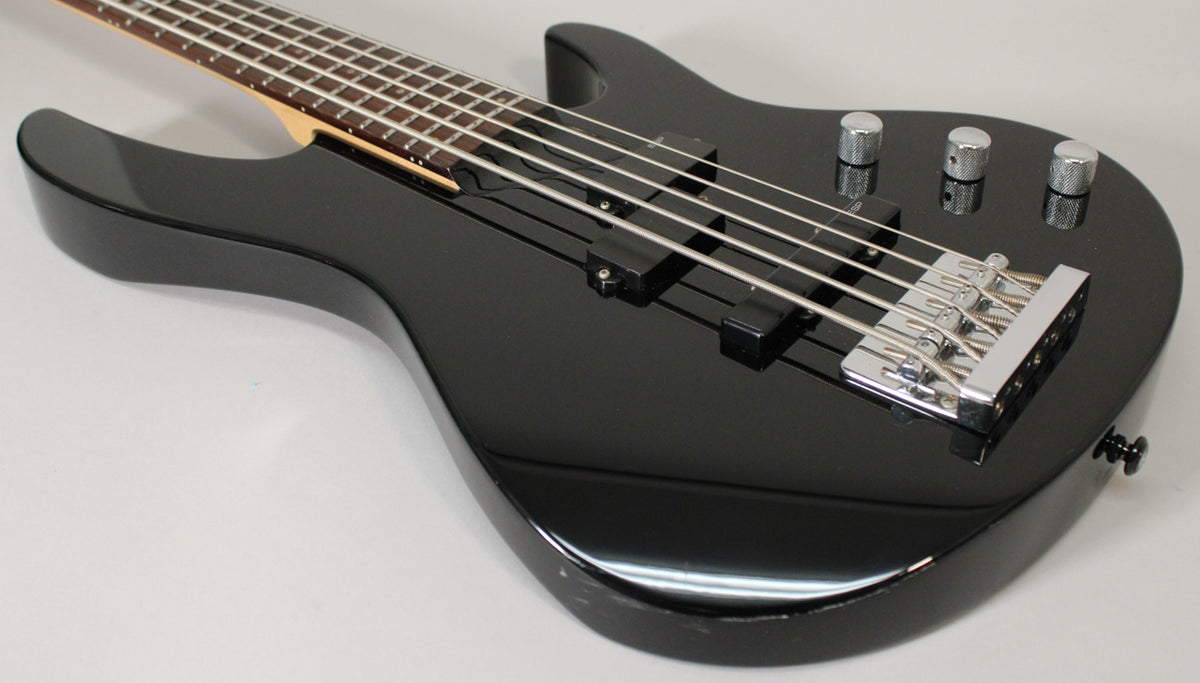 The glossy black 5-string bass is angled from the upper left, showing its two pickups, chrome bridge, and three control knobs.