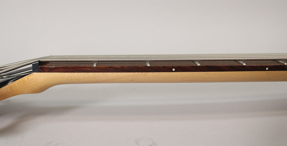 Close-up of the bass neck from the side, showing the light wood neck, dark fretboard, silver strings, and white dot markers.
