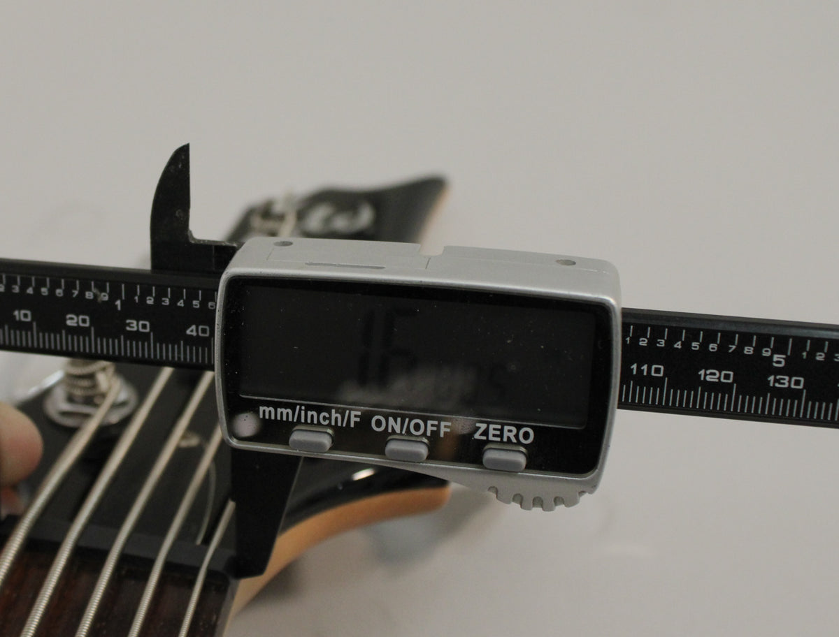 A digital caliper measures the nut width of the black bass guitar, showing 1.65 inches.