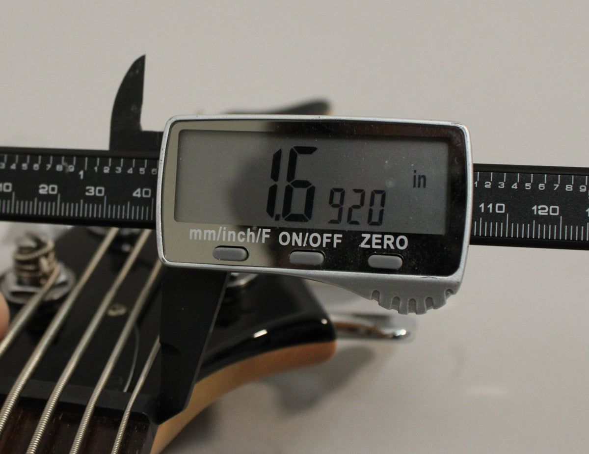 A digital caliper measures the nut width of the black bass guitar's headstock, displaying "1.6920 in".