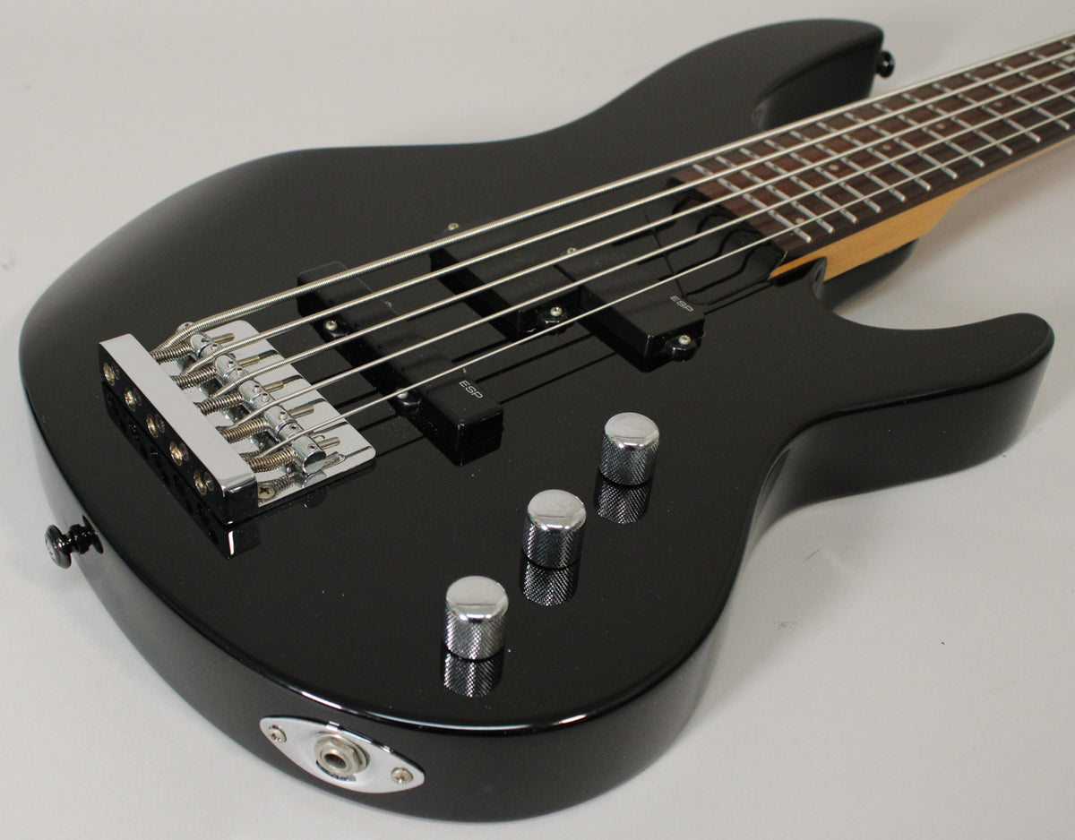 Close-up of a glossy black 5-string bass, angled from the lower bout, showing the chrome bridge, two black pickups, and three knurled chrome knobs.