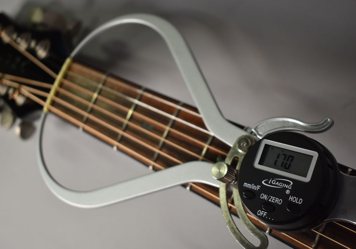 A close-up of the guitar's fretboard and headstock, with a digital caliper measuring the string height at the first fret.