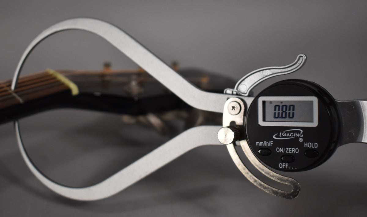 A digital caliper measures the black guitar's neck, showing 0.00 on its screen, with the fretboard and nut visible.