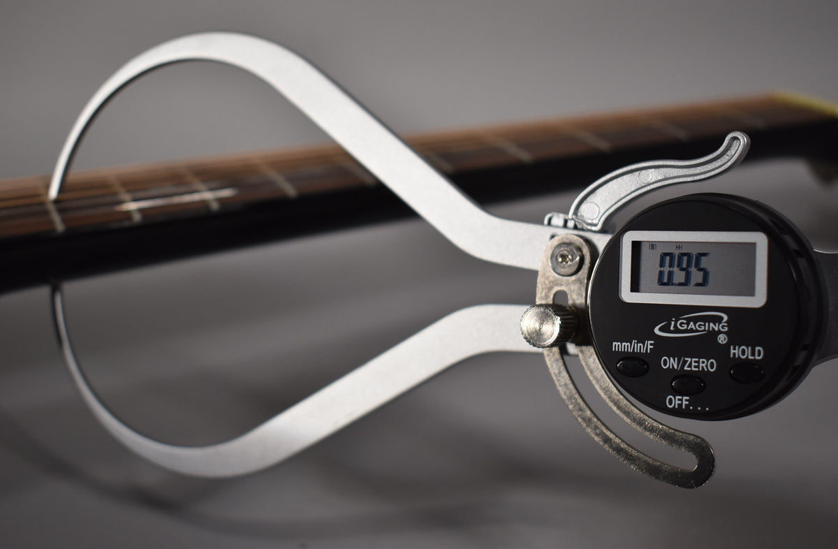 A digital caliper measures the string height at the 12th fret of the black acoustic guitar's dark fretboard.