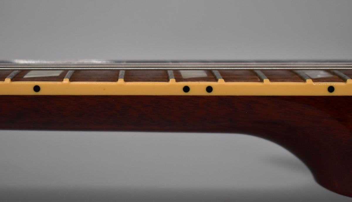 The guitar's dark brown neck is seen from the side, showing cream binding, black side dots, and fretboard with trapezoid inlays.