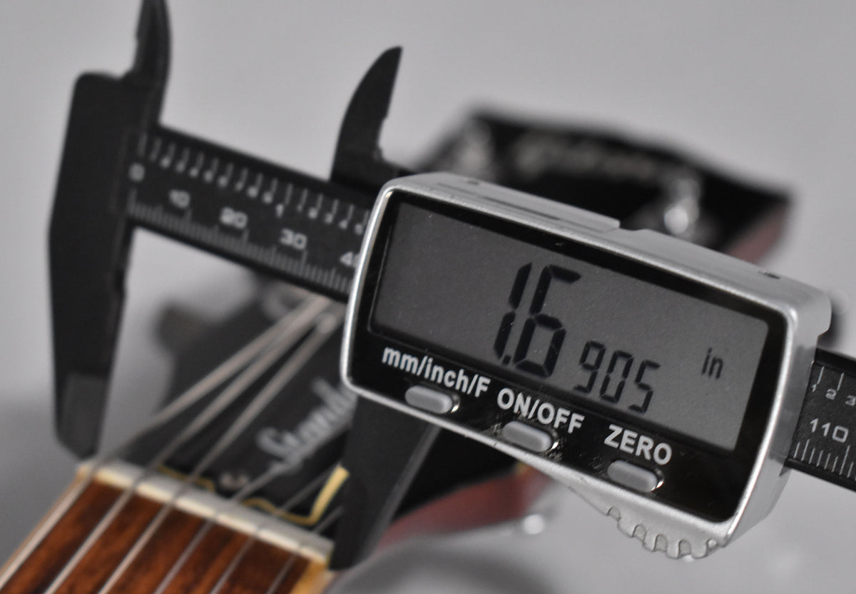 A digital caliper measures the nut width of the guitar neck, showing 1.6905 inches, with the dark headstock visible behind.