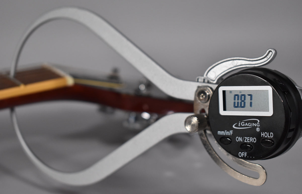 A digital caliper measures the neck thickness at the first fret, displaying 0.87 inches, with the fretboard and headstock out of focus.