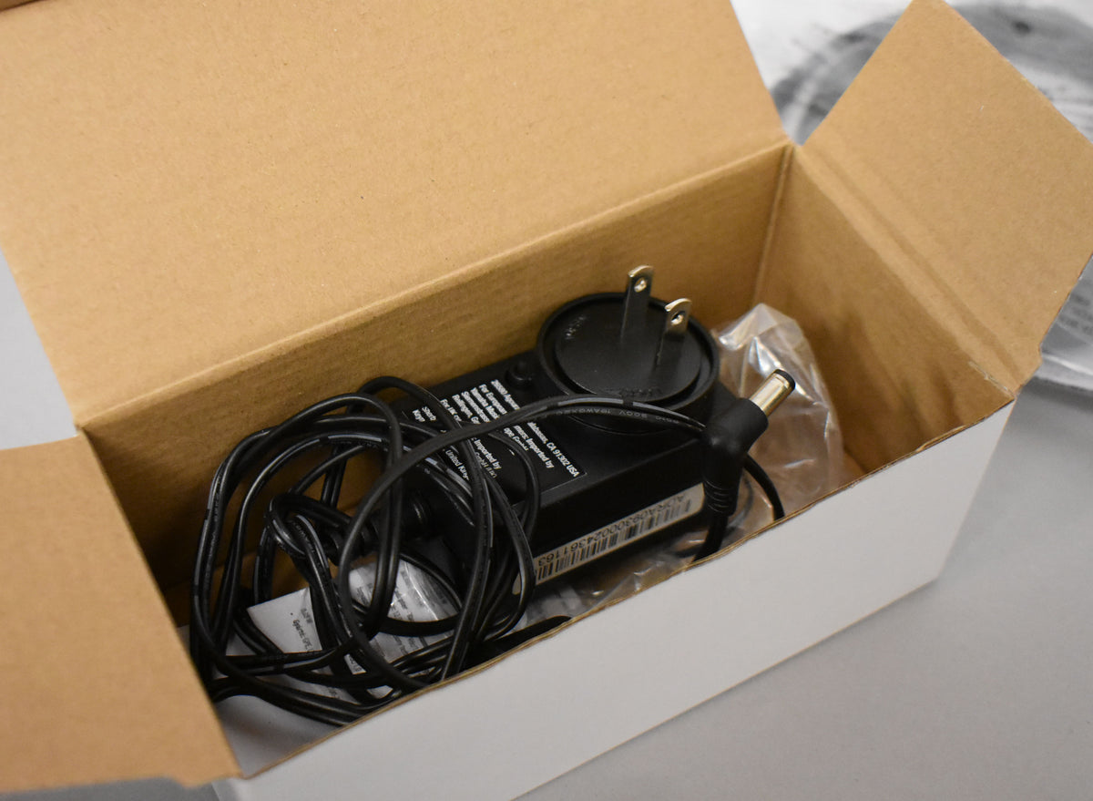 A black power adapter with a coiled cord and a two-prong plug rests inside a white and brown cardboard box.