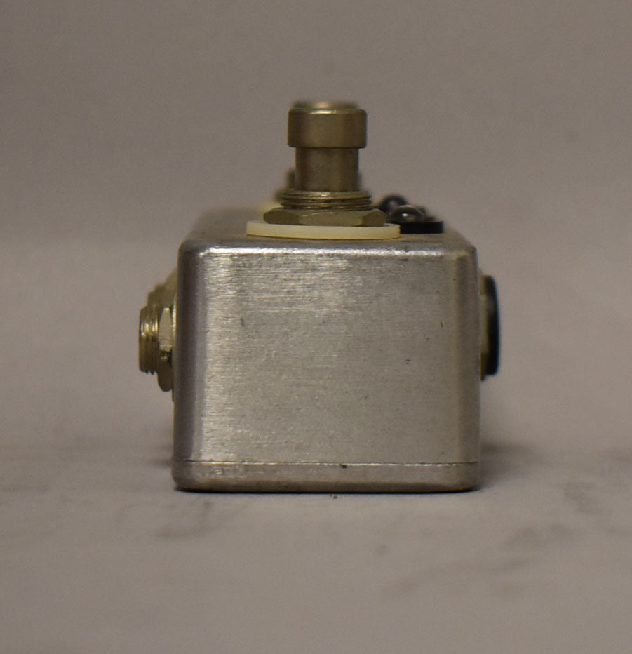 The brushed aluminum pedal is viewed from the side, showing a brass footswitch and a jack on the left.