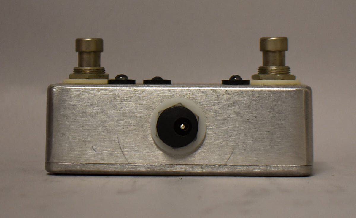 A brushed metal guitar pedal with two footswitches, three small black indicator lights, and a power input jack.