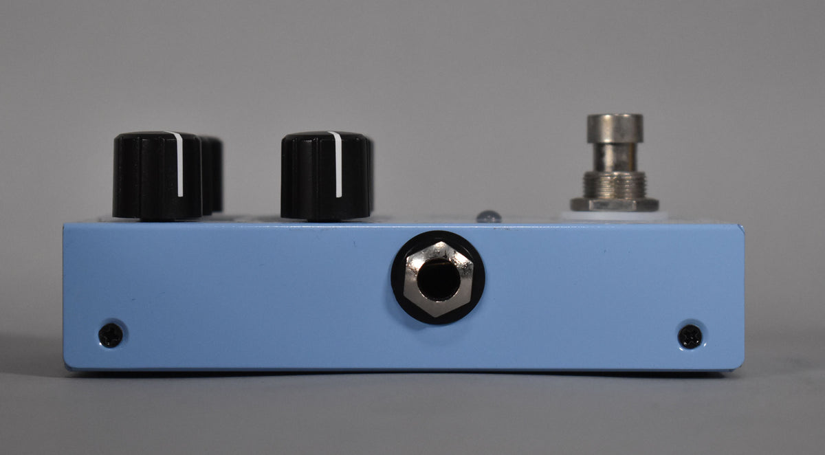 A light blue guitar pedal with two black knobs, a silver input jack, and a silver footswitch on a gray background.