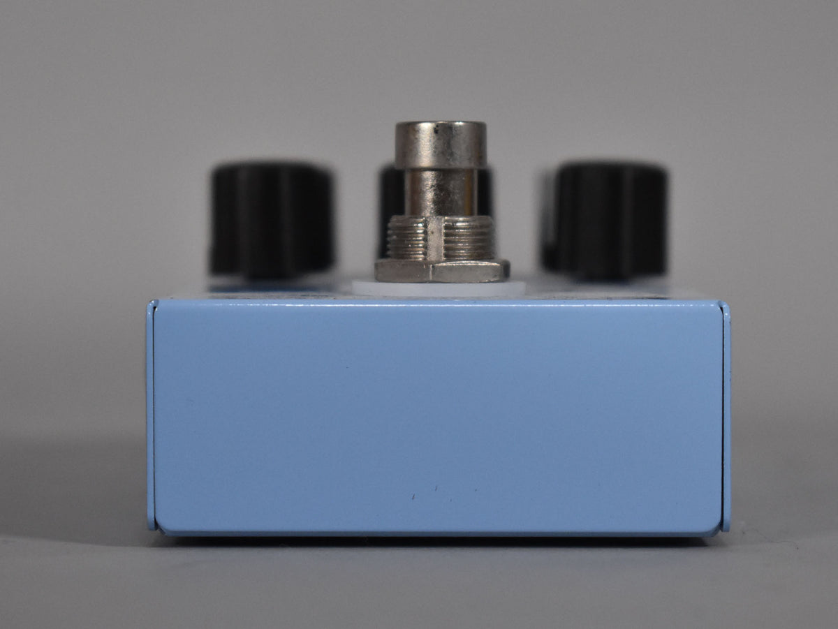 The light blue pedal is viewed from the front, showing a silver jack and two black control knobs on top.