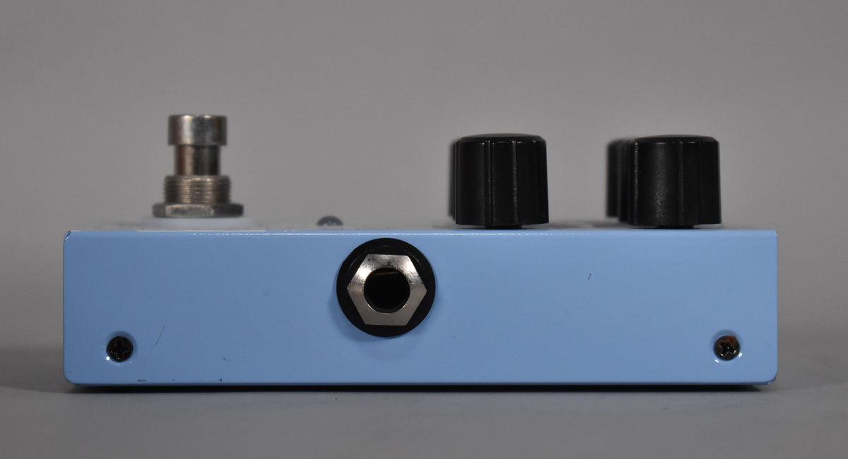 The light blue pedal is viewed from the side, showing a chrome input jack, two black knobs, and a silver footswitch.