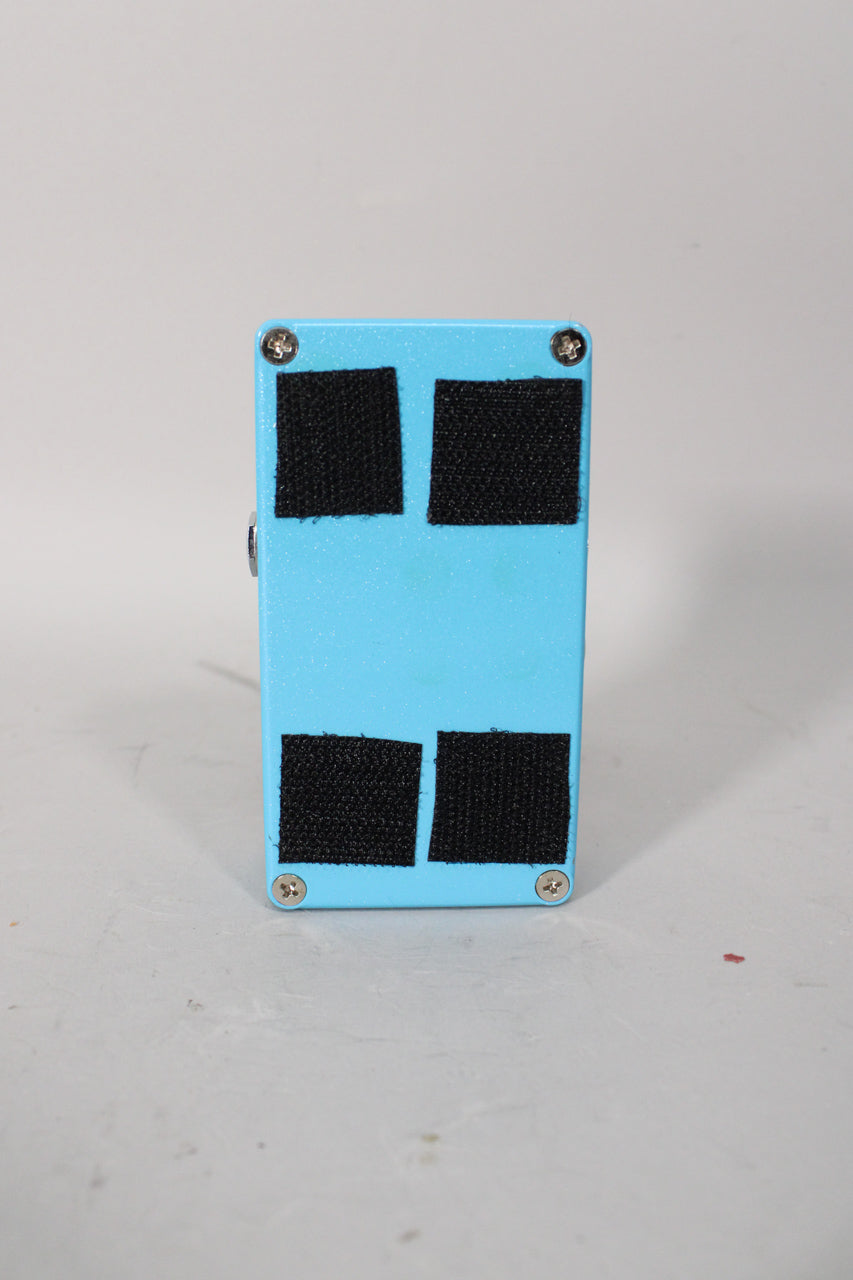 The light blue pedal is seen from the back, showing four black velcro squares and four silver screws on the textured finish.