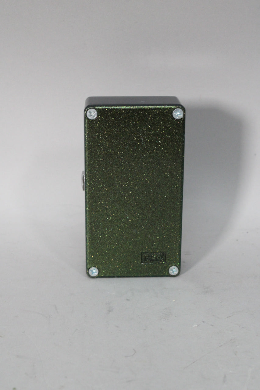The back of the dark green glitter-finish pedal is visible, secured by four silver screws, with a small black rectangular logo near the bottom.