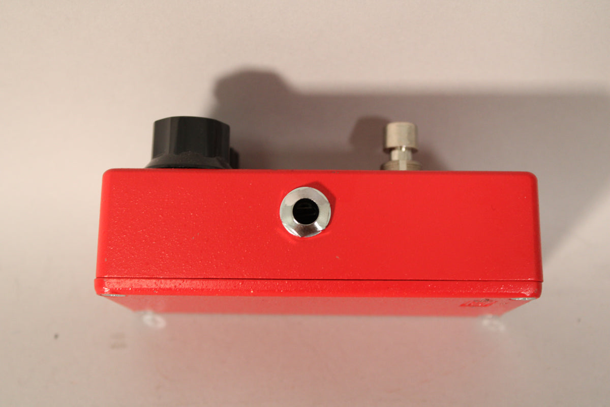 The red pedal is viewed from the side, showing a black knob, a silver input jack, and a silver output jack.