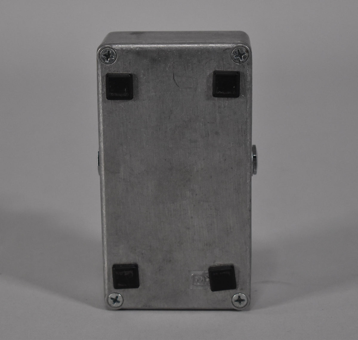 The silver metal pedal is viewed from the bottom, showing four black rubber feet and four Phillips head screws.