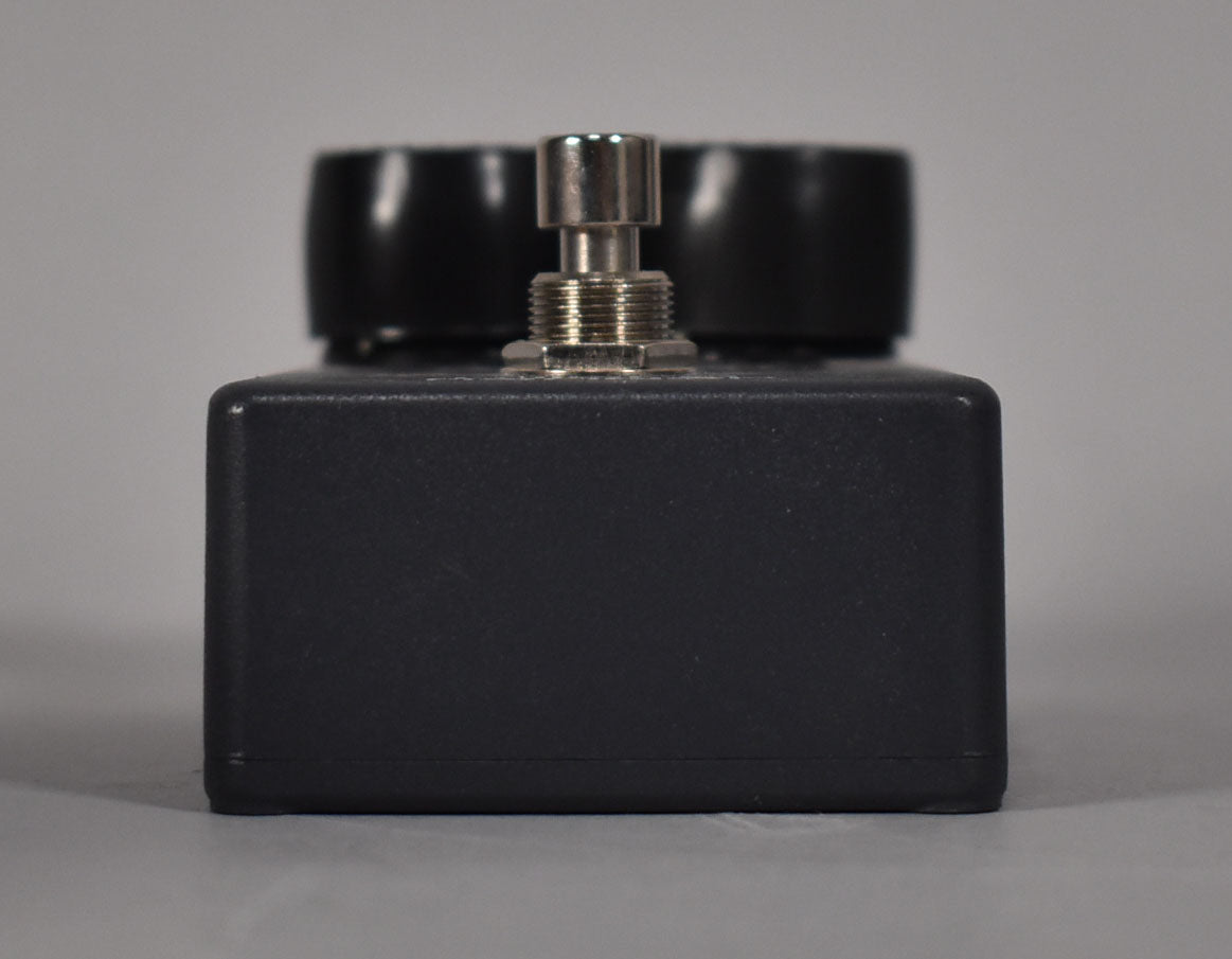 The dark gray pedal is viewed from the side, revealing a chrome footswitch and two black control knobs behind it.