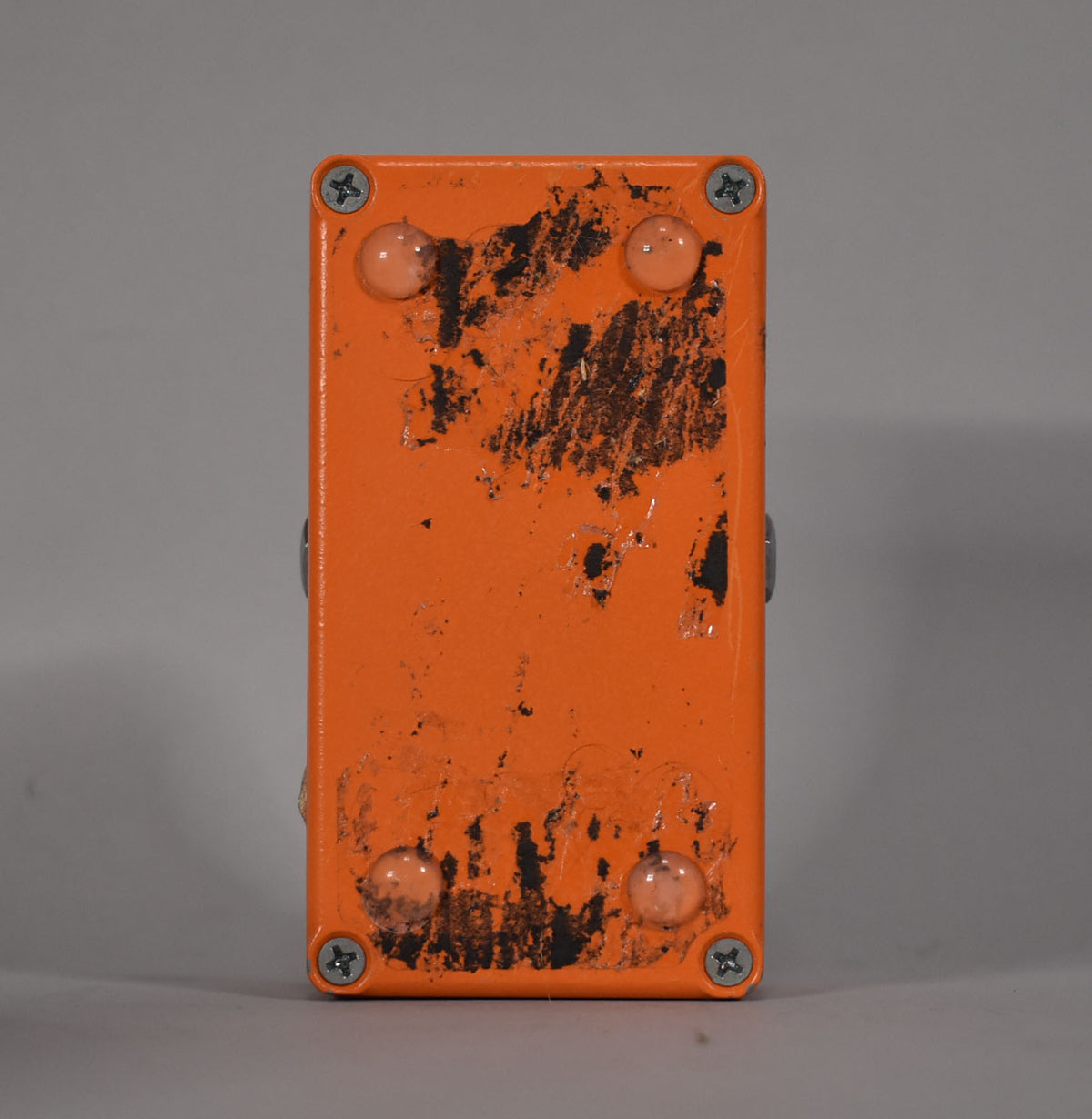 The orange pedal is viewed from the bottom, showing four silver screws, four clear rubber feet, and heavy black scuffing and wear.