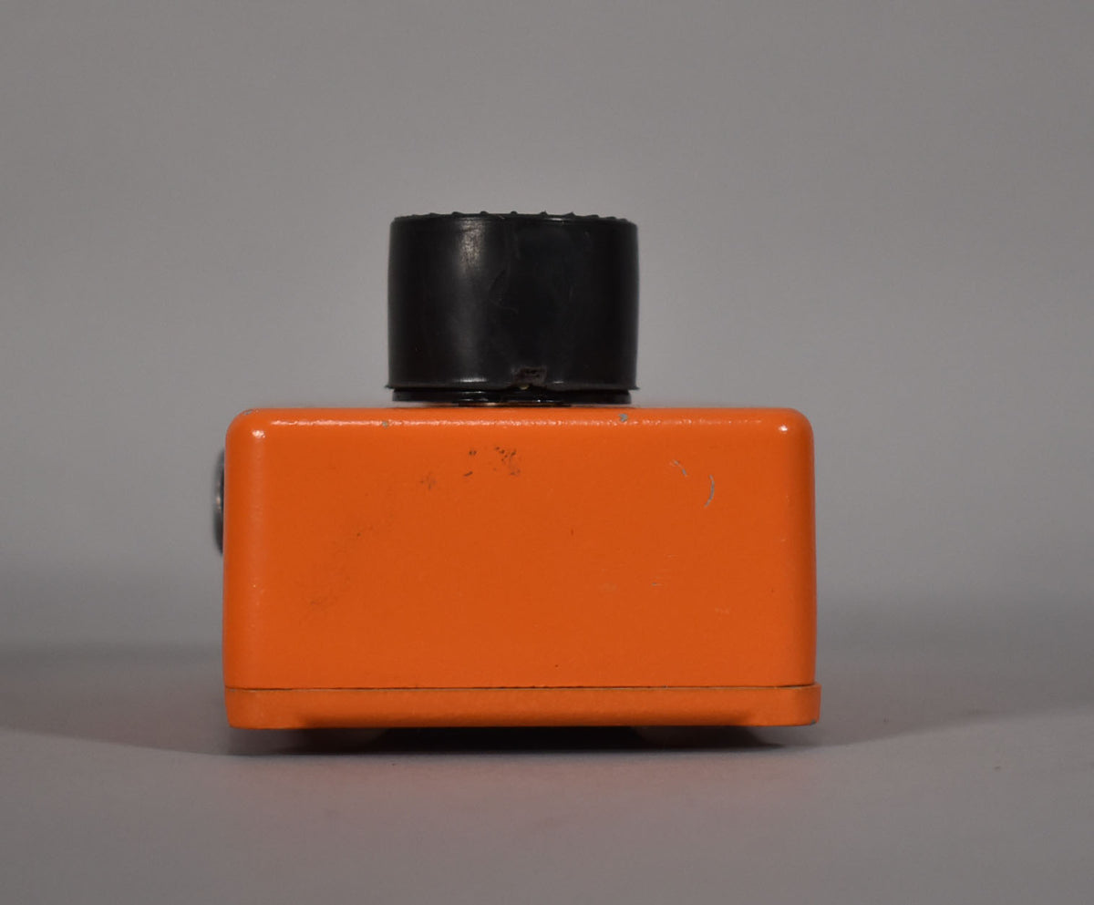 The orange pedal is viewed from the right side, showing a black knob on top and minor scuffs on its textured finish.