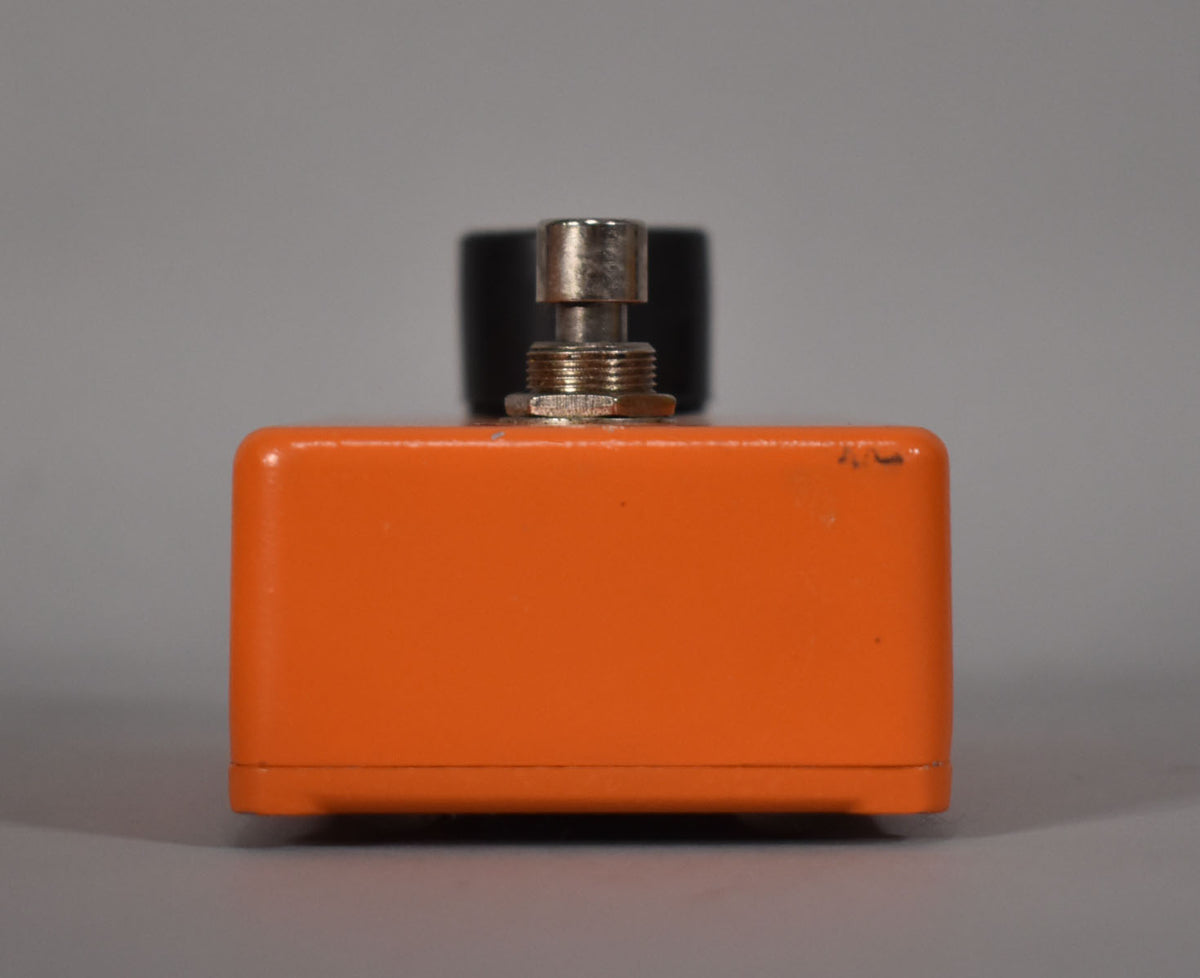 The orange pedal is viewed from the side, showing a silver footswitch and minor scuffs on the top right edge.
