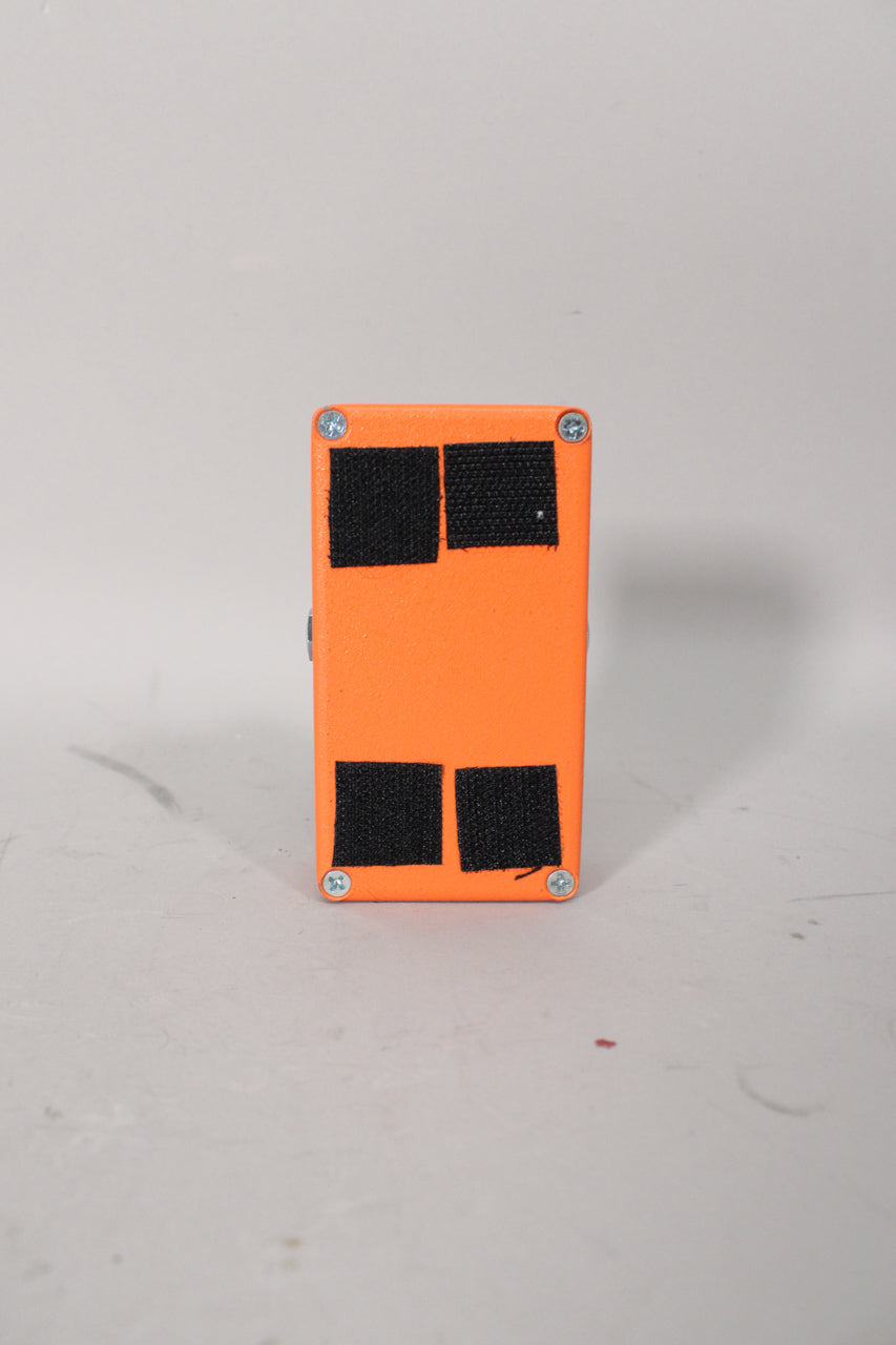 The orange pedal is seen from the bottom, revealing four silver screws and four rectangular black velcro pads.