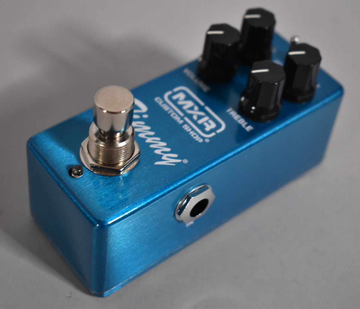 A metallic blue mini pedal, angled from the front right, showing a silver footswitch, four black control knobs, and a side input jack.