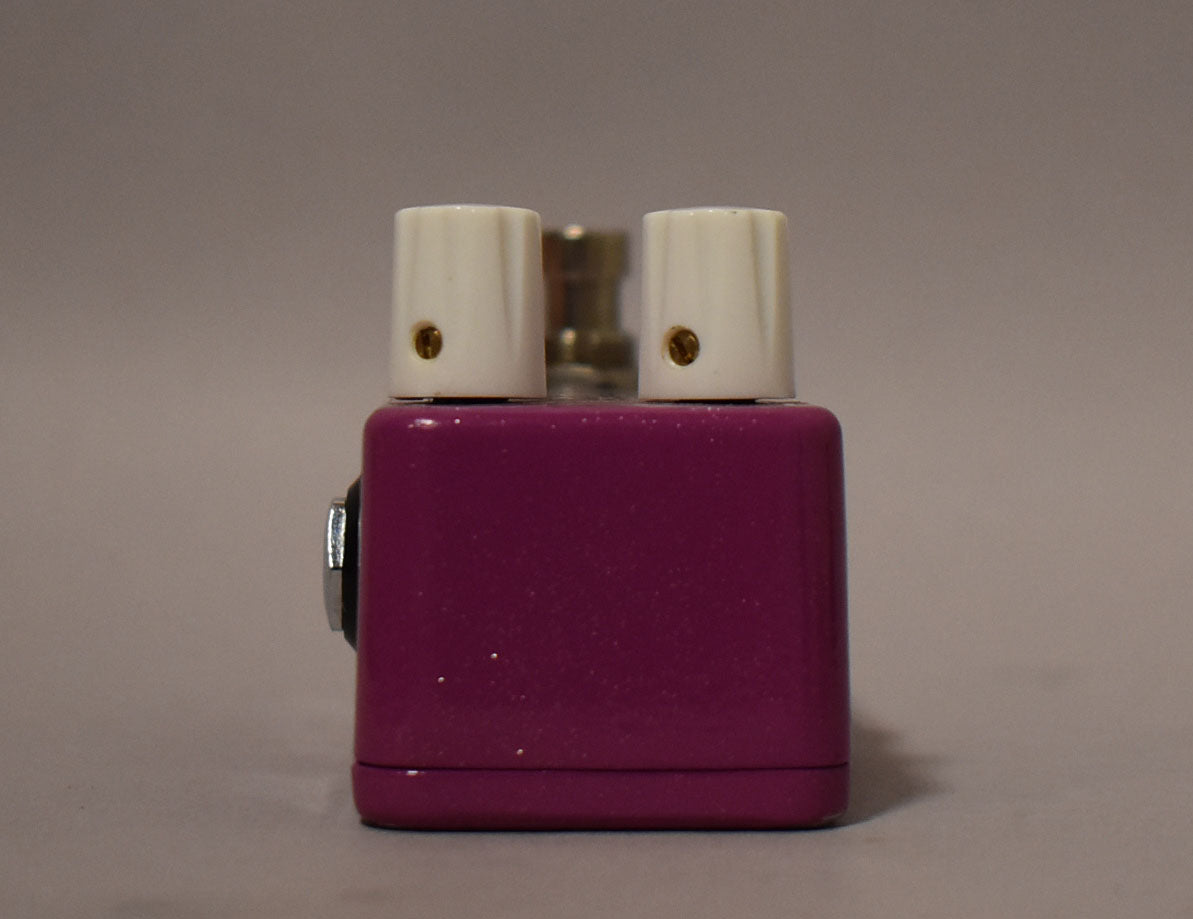 Front view of a shiny purple pedal with two white knobs on top, each with a gold indicator dot.
