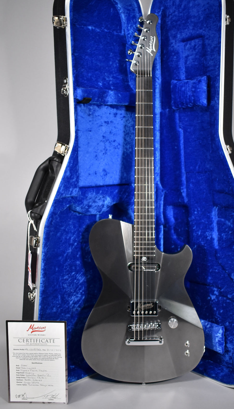 A dark gray electric guitar with a faceted body and chrome hardware stands upright in a blue velvet-lined hard case.