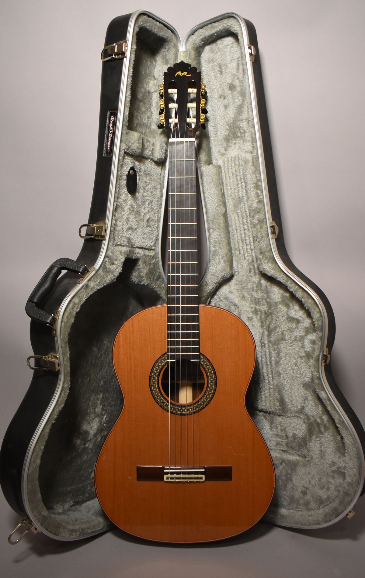 Full-front view of a classical guitar with a light brown body, dark fretboard, and gold tuners, standing in its open hard case.
