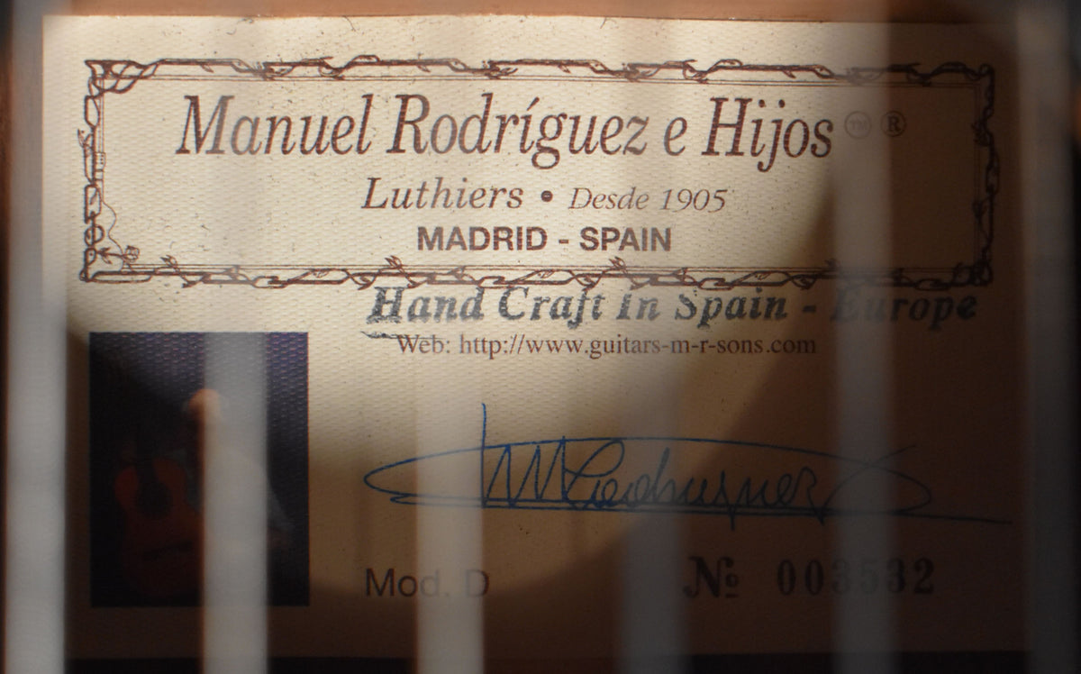 The guitar's interior label, framed by dark wood, shows a decorative border, text, a blue signature, and a small square image.