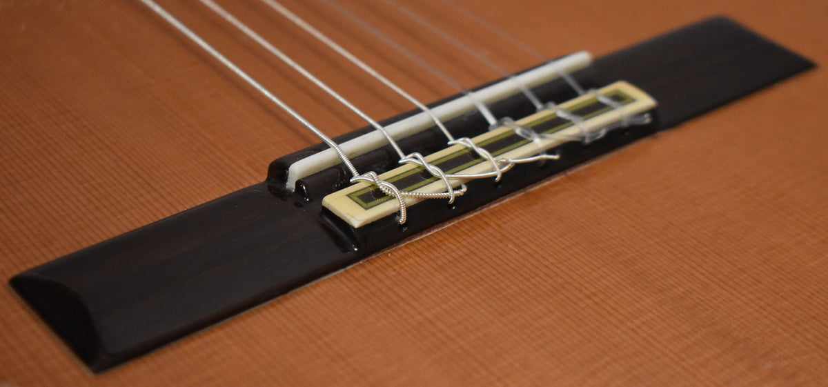 Close-up, high-angle view of the classical guitar's bridge, showing the dark wood, light saddle, and tied nylon strings.