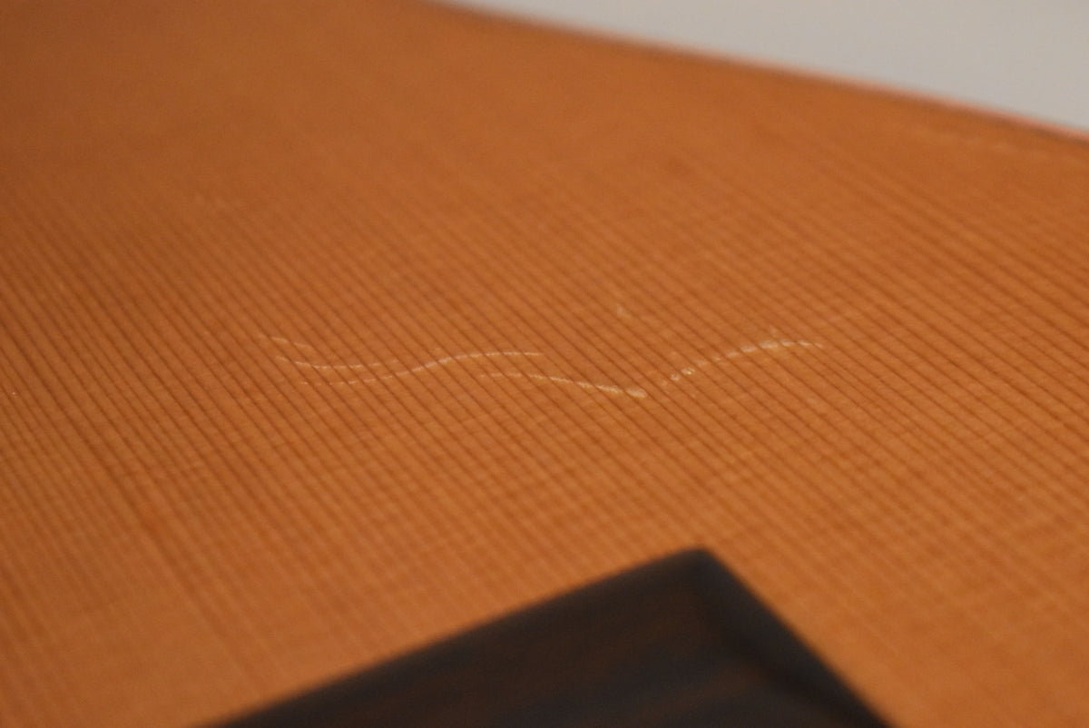 Close-up of the guitar's light brown top, showing a wavy scratch across the grain and the dark corner of the bridge.