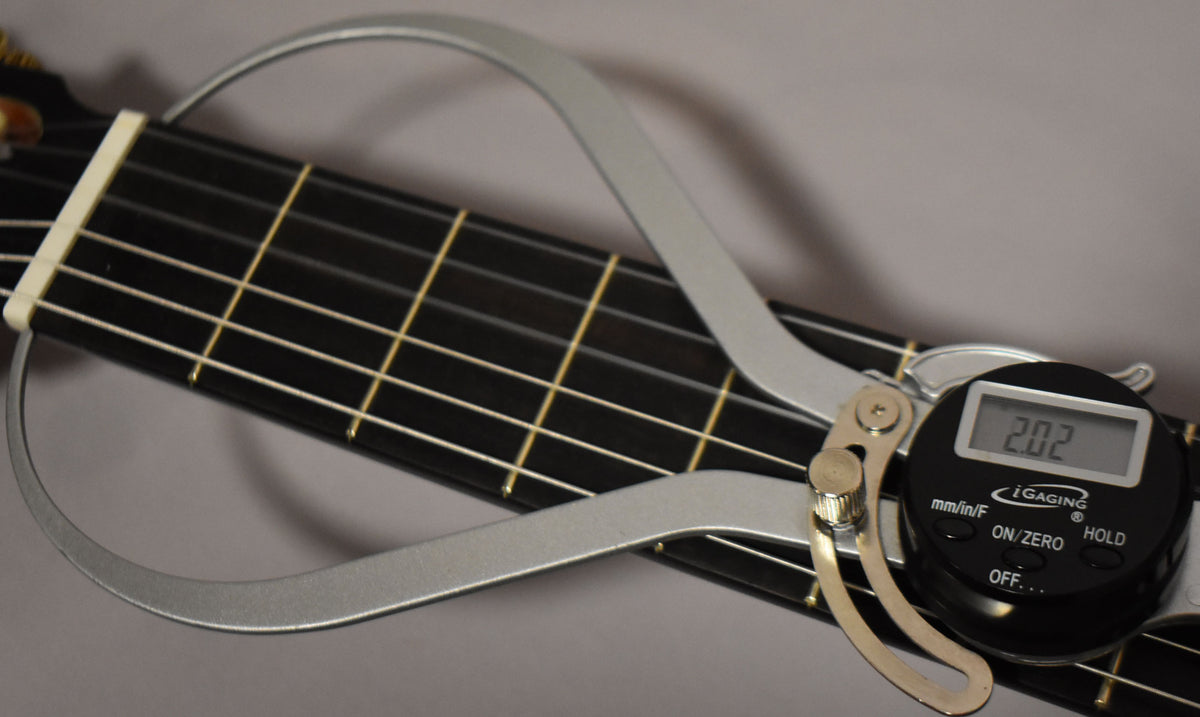 Close-up of the guitar's dark fretboard and light frets, with a silver digital caliper measuring the string height at 2.02.