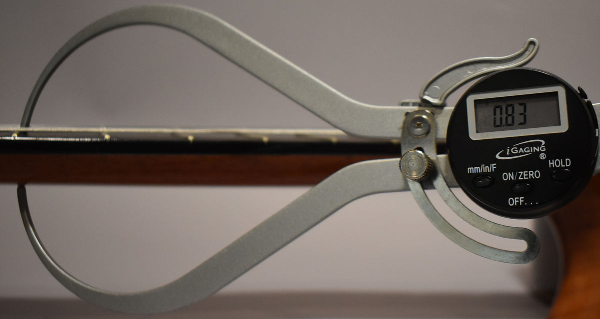 A digital caliper measures the guitar's dark brown neck, showing a reading of 0.83 on its black display.