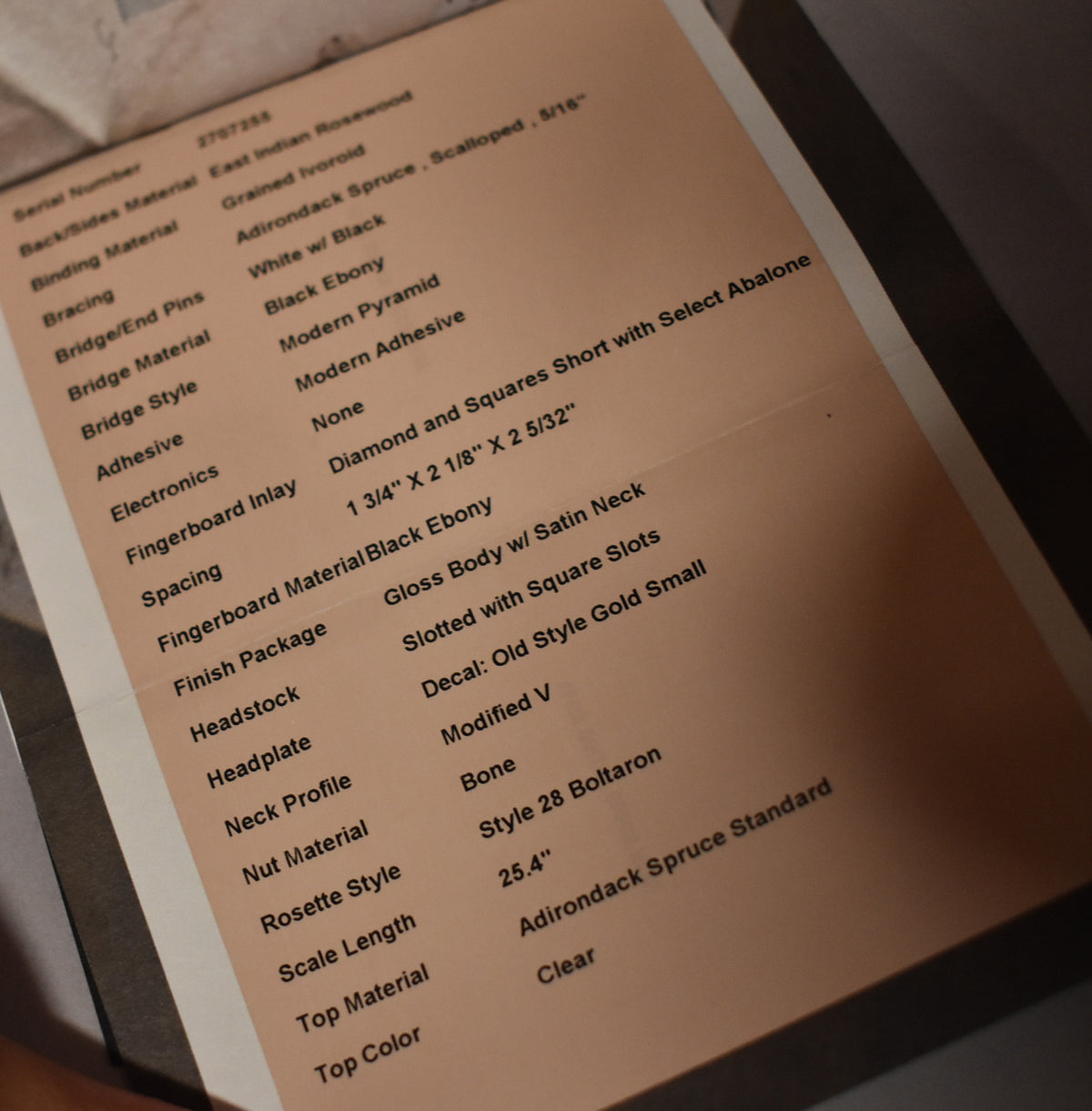 A close-up, angled view of a cream-colored spec sheet with black text, listing guitar features like "East Indian Rosewood" and "Gloss Body w/ Satin Neck."