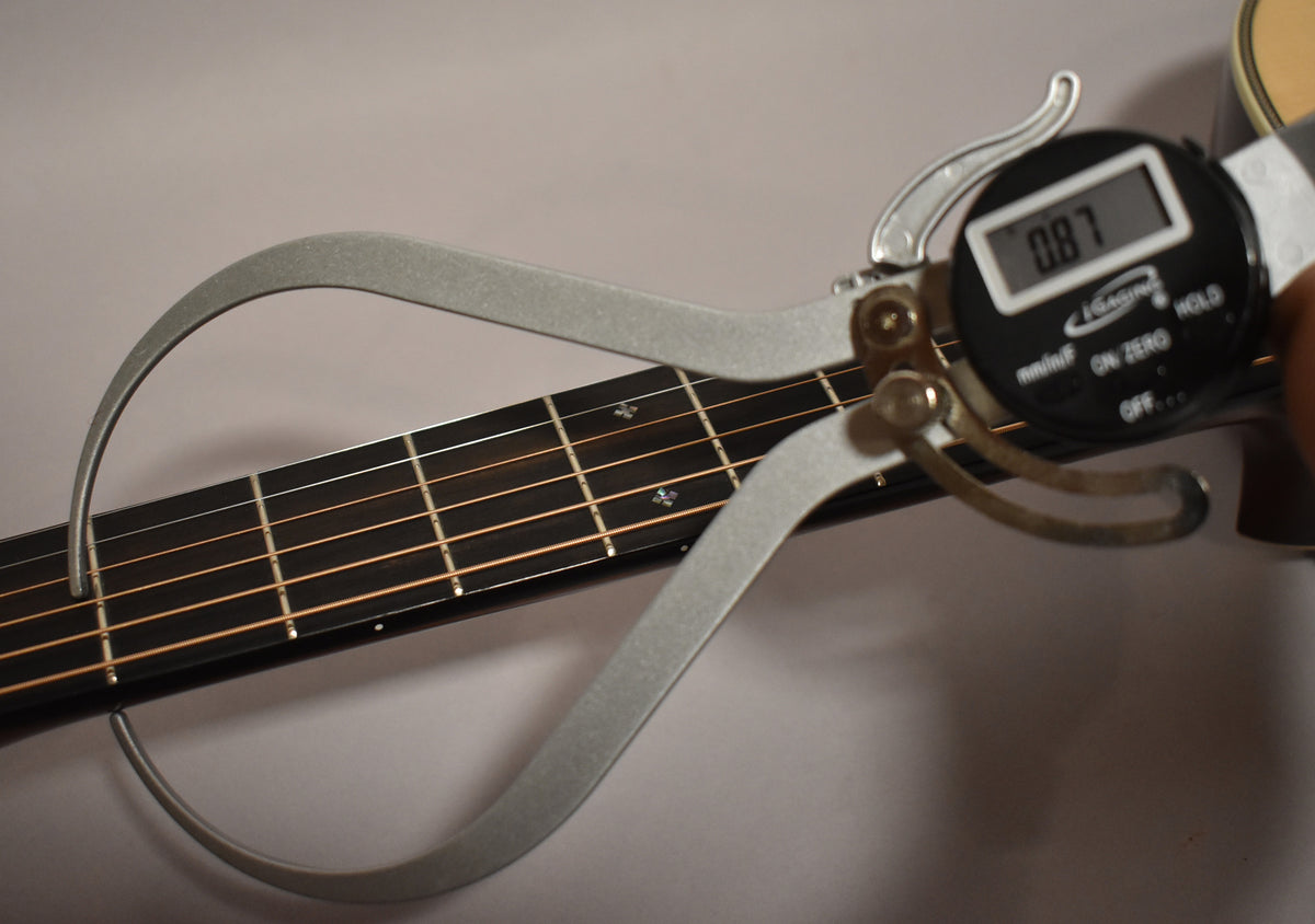 A digital caliper measures the guitar's fretboard at 0.87 inches, showing the dark fretboard and light strings.