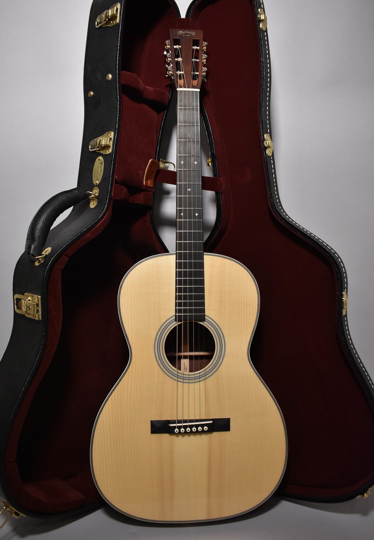 Full-front view of a light wood acoustic guitar with a dark fretboard and bridge, standing upright in its open, dark red-lined case.
