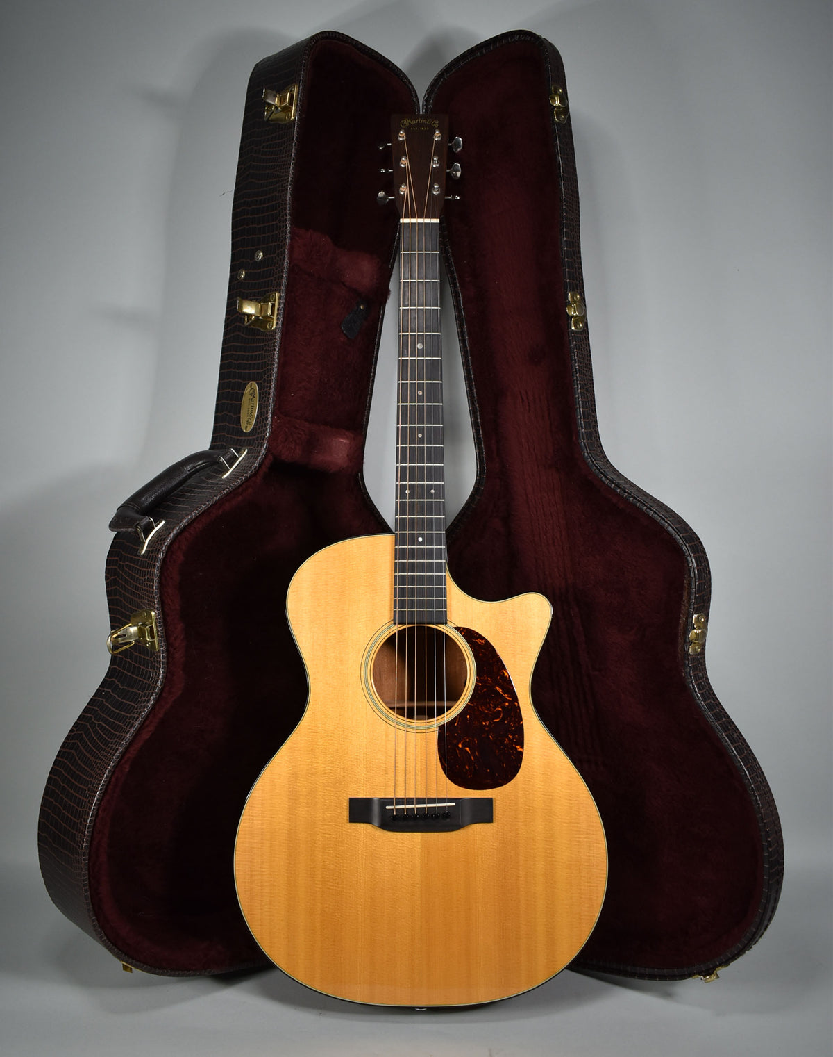 Full-length view of a light wood acoustic guitar with a cutaway body, standing upright in front of its open, dark brown hard shell case.